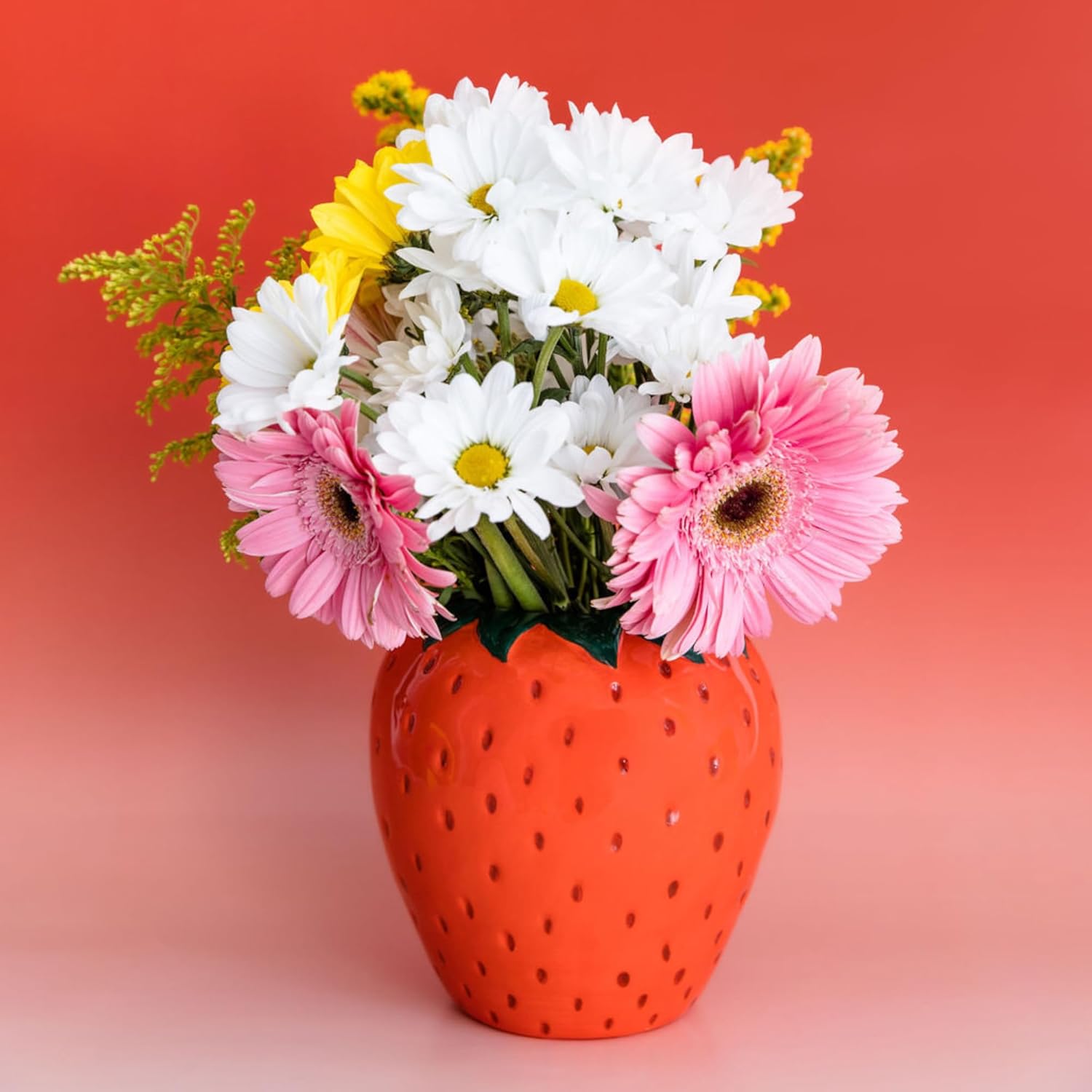 Bright red ceramic vase shaped like a strawberry, filled with colourful daisies and pink gerbera flowers.