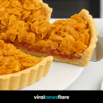 A close-up of a traditional British cornflake tart with a layer of raspberry jam, served on a white plate with a slice being lifted.
