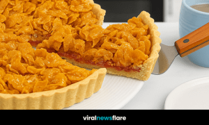 A close-up of a traditional British cornflake tart with a layer of raspberry jam, served on a white plate with a slice being lifted.